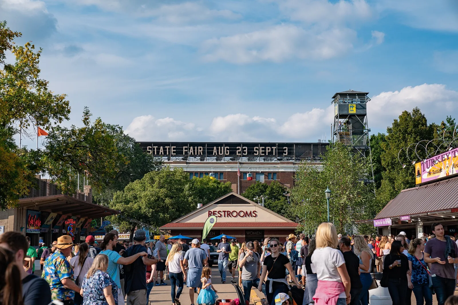 A dense, happy crowd streaming past food stands under the Minnesota State Fair gateway sign on a bright day